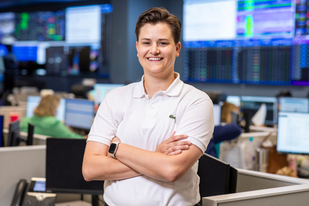Children's Mercy employee in white polo shirt with arms crossed and smiling in room filled with monitors on the wall in the background. 