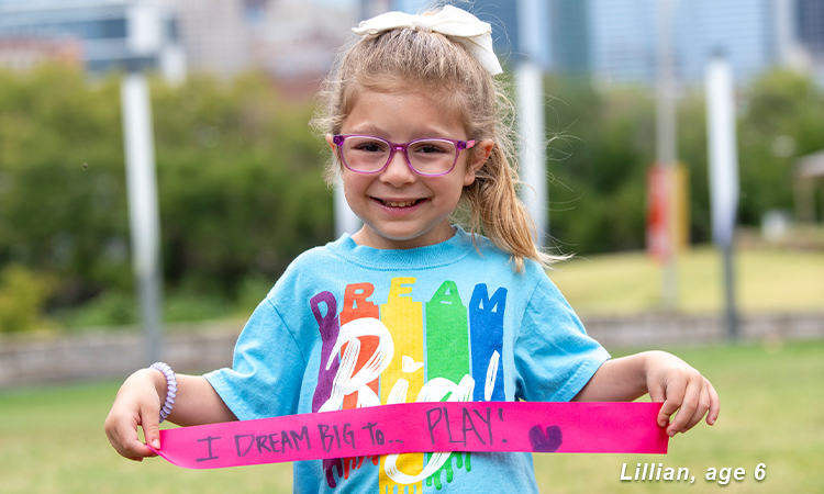 Lillian, age 6. She is smiling outside while holding a ribbon that reads: I dream big to play! 