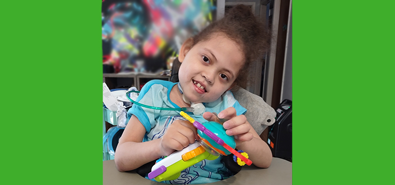 A photo on a green background. In the photo a young girl with a breathing tube sits and plays with a toy.