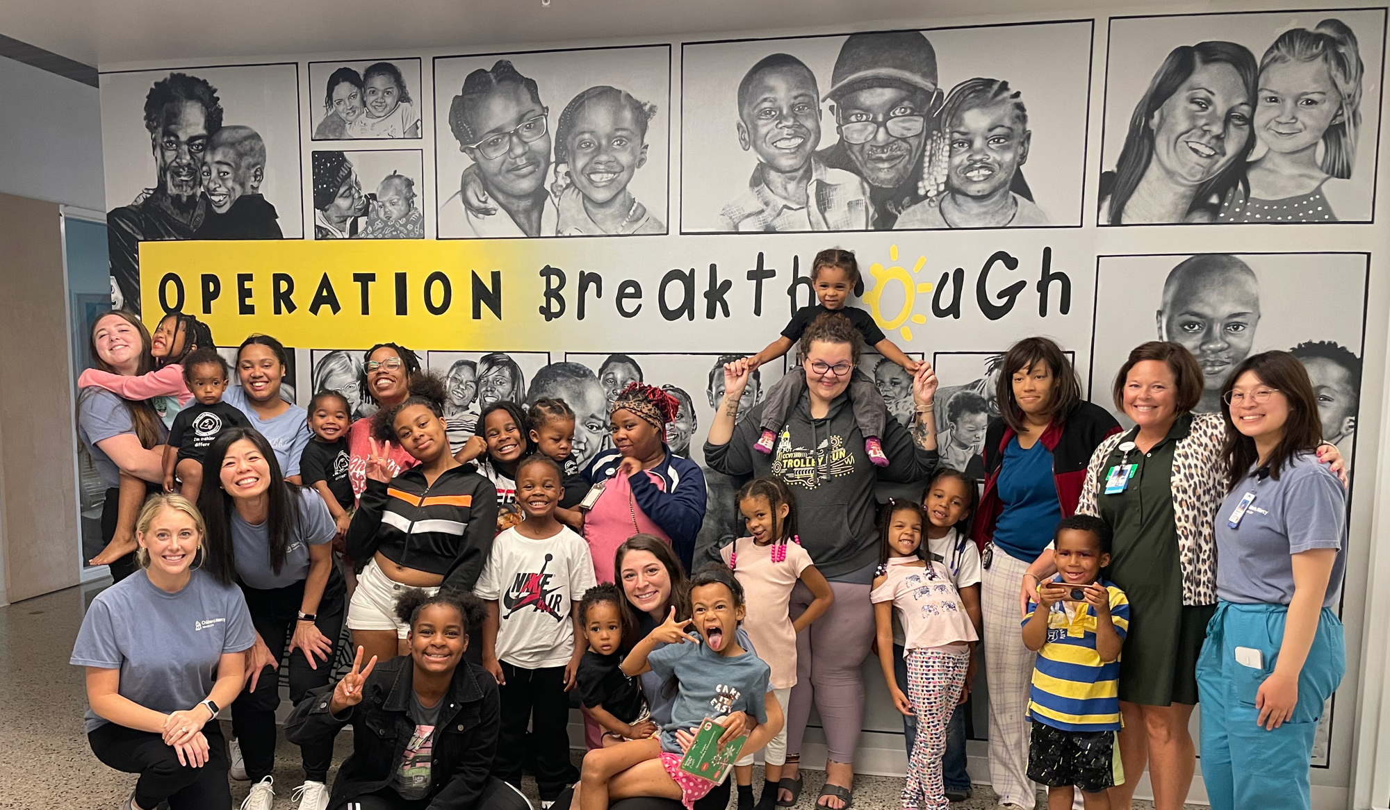 A large group of women and children pose in front of a wall mural that shows black and white drawings of people. The wall mural also says Operation Breakthrough on it.