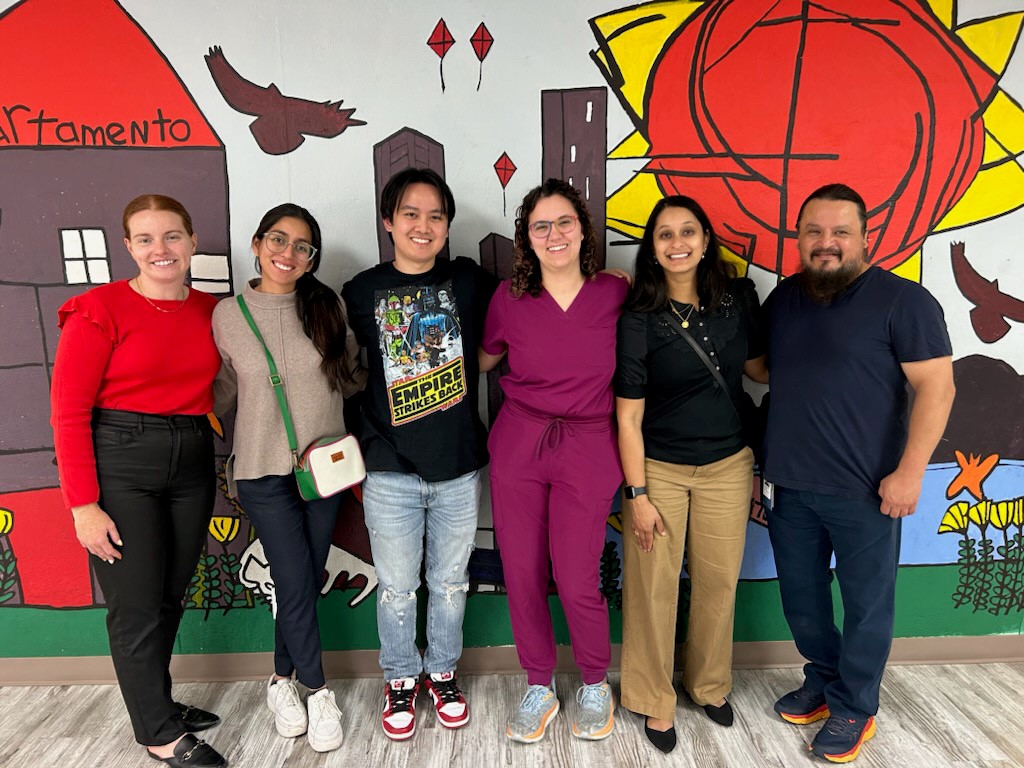 Six people stand together in front of a colorful mural featuring buildings, birds, and a large sun, posing for a group photo indoors.