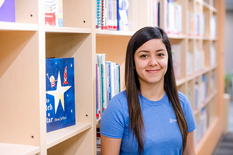 Person in a blue Children’s Mercy T-shirt standing beside bookshelves in a library or learning center. Books and educational materials line the shelves in the background.