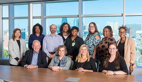 12 people, men and women, pose in a conference room. Four of the people sit at a table while the rest stand.