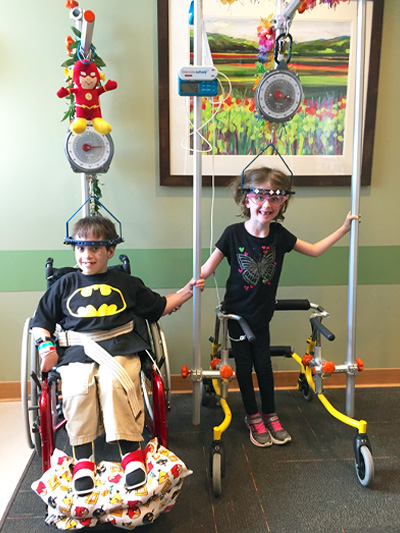 Two children using body-weight support harness systems during pediatric rehabilitation therapy, with adaptive mobility equipment visible