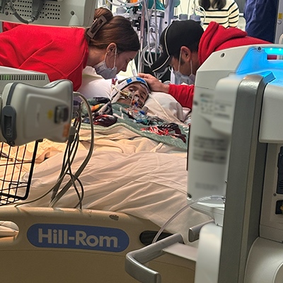 A small boy is in a hospital bed. He has huge tubes attached to his mouth and wires everywhere. His eyes are shut. There’s a colorful quilt covering up half his body. His mother leans over him, and his dad is standing behind, looking at something out of the frame. 