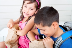 Two children are sitting side by side, each holding a brown paper bag. The child on the left is wearing a pink dress and a pink headband, while the child on the right is wearing a blue shirt and has a backpack on. They appear to be looking into their bags. The background consists of white horizontal siding.