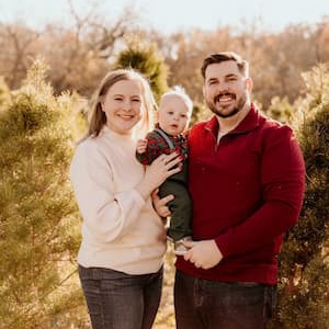 Family portrait taken outdoors featuring two adults and a young child standing together among evergreen trees. One adult is holding the child, and all three are facing the camera. The photo is lit by warm natural sunlight with trees and soft-focus foliage in the background.