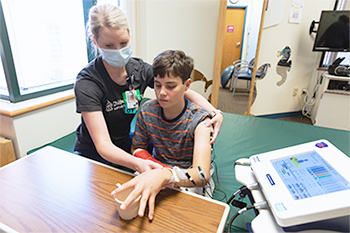 Child participating in a rehabilitation therapy session while a therapist assists with hand and arm movements using a computerized assessment device