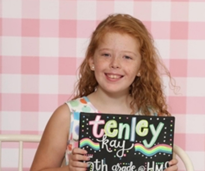 School-age child holding a chalkboard-style first-day-of-school sign while seated in front of a pink checkered backdrop.