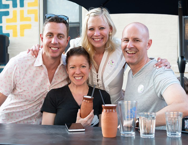 Group of four adults gathered at an outdoor patio table, with drinks and personal items in front of them, posing for a photo.