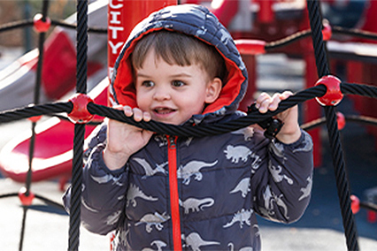 Young child climbing on a rope playground structure while wearing a hooded jacket with a dinosaur pattern.