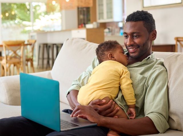 A Black father sits on a couch holding a sleeping baby while using a laptop at home.