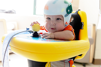 Infant wearing a therapy helmet and seated in a supportive mobility device while interacting with a toy