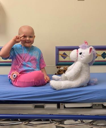 Child sitting on a hospital bed beside a large stuffed unicorn.