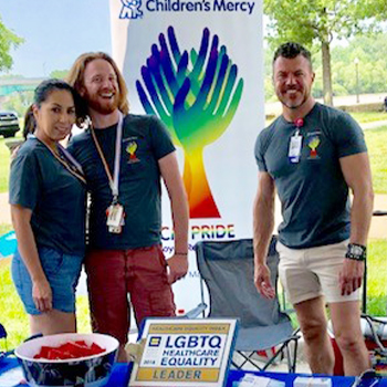 Three people standing at a Children's Mercy Pride event booth outdoors, with a rainbow-colored hands display and signage recognizing LGBTQ+ Healthcare Equality.