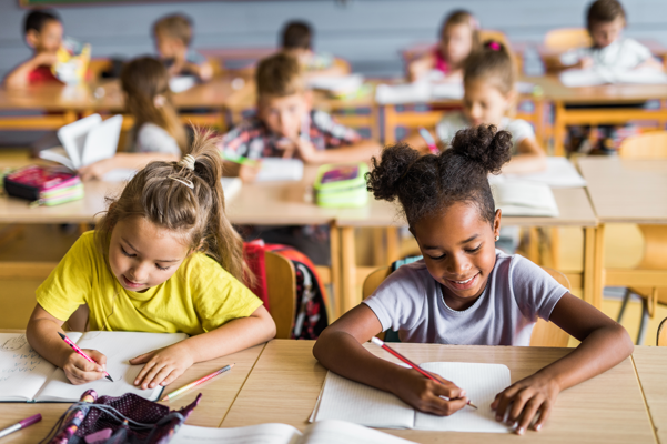 Students seated at desks in a classroom, working on writing assignments. Two students in the foreground are focused on drawing or writing in notebooks, while additional students are seated at desks in the background.