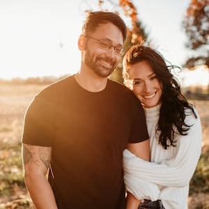 Outdoor portrait of two people standing together in a sunlit field. One person is wearing a dark short-sleeve shirt and glasses, and the other is wearing a light-colored sweater. Trees and warm golden-hour lighting are visible in the background.