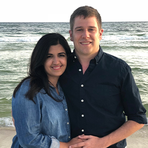 Two people standing together on a beach with ocean waves in the background. One person is wearing a light blue button-up shirt, and the other is wearing a dark button-up shirt. The pair is posed close together near the shoreline.