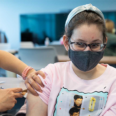 A young woman wearing a face mask receives a vaccine in her arm.