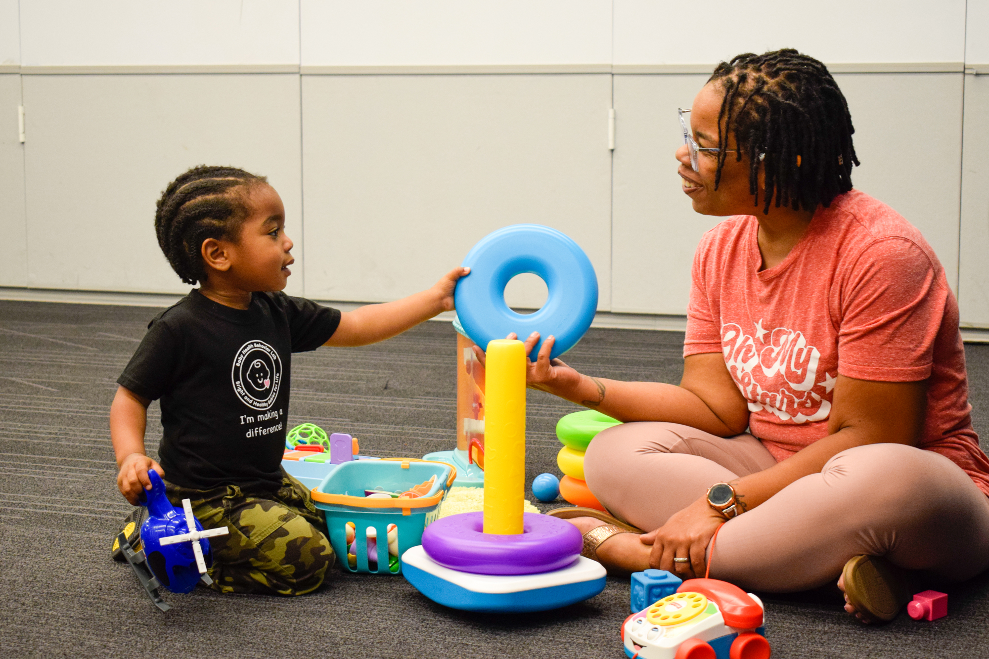 A mother and her son sit on the floor playing with toys. He hands her a toy.
