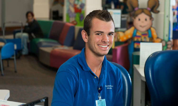 Volunteer seated in a pediatric hospital waiting area, wearing a Children's Mercy volunteer shirt and identification badge.