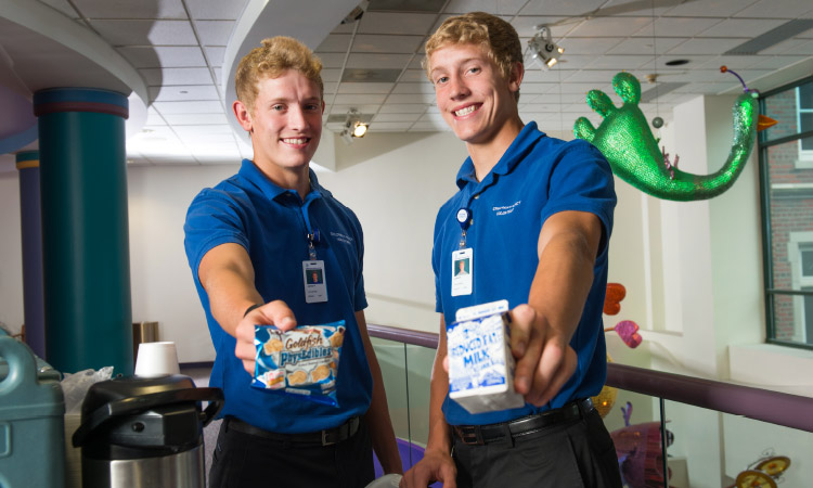 Two Children's Mercy volunteers holding cartons of milk and packaged snacks while standing in a pediatric hospital unit.