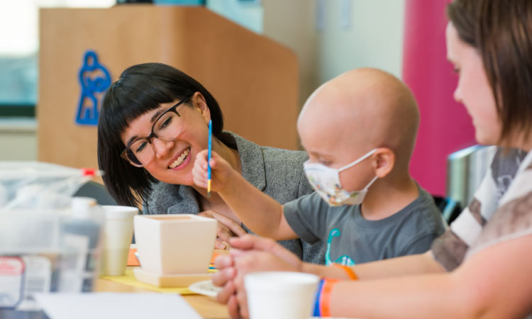 Child participating in an arts-and-crafts activity with support from two adults at a table in a pediatric care setting.