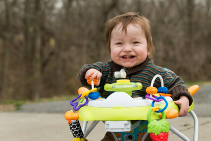 Young child seated in a baby activity walker outdoors, interacting with colorful attached toys, with trees visible in the background.