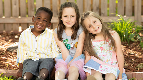 Three children sitting together outdoors in a garden setting with a wooden fence and landscaping in the background.