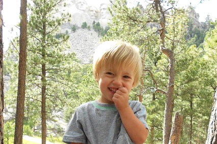 Young child standing among pine trees in a mountain landscape, with a rocky cliff visible in the distance
