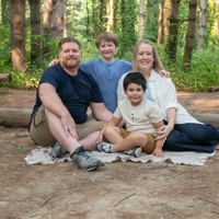 photo of family of four posing outside while sitting on the ground near a log and under forest trees