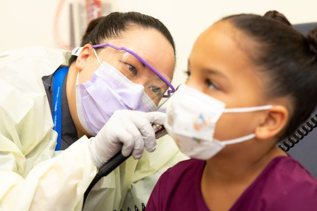 Clinician wearing protective mask, face shield, and gloves examines a child’s ear with an otoscope during a medical visit.