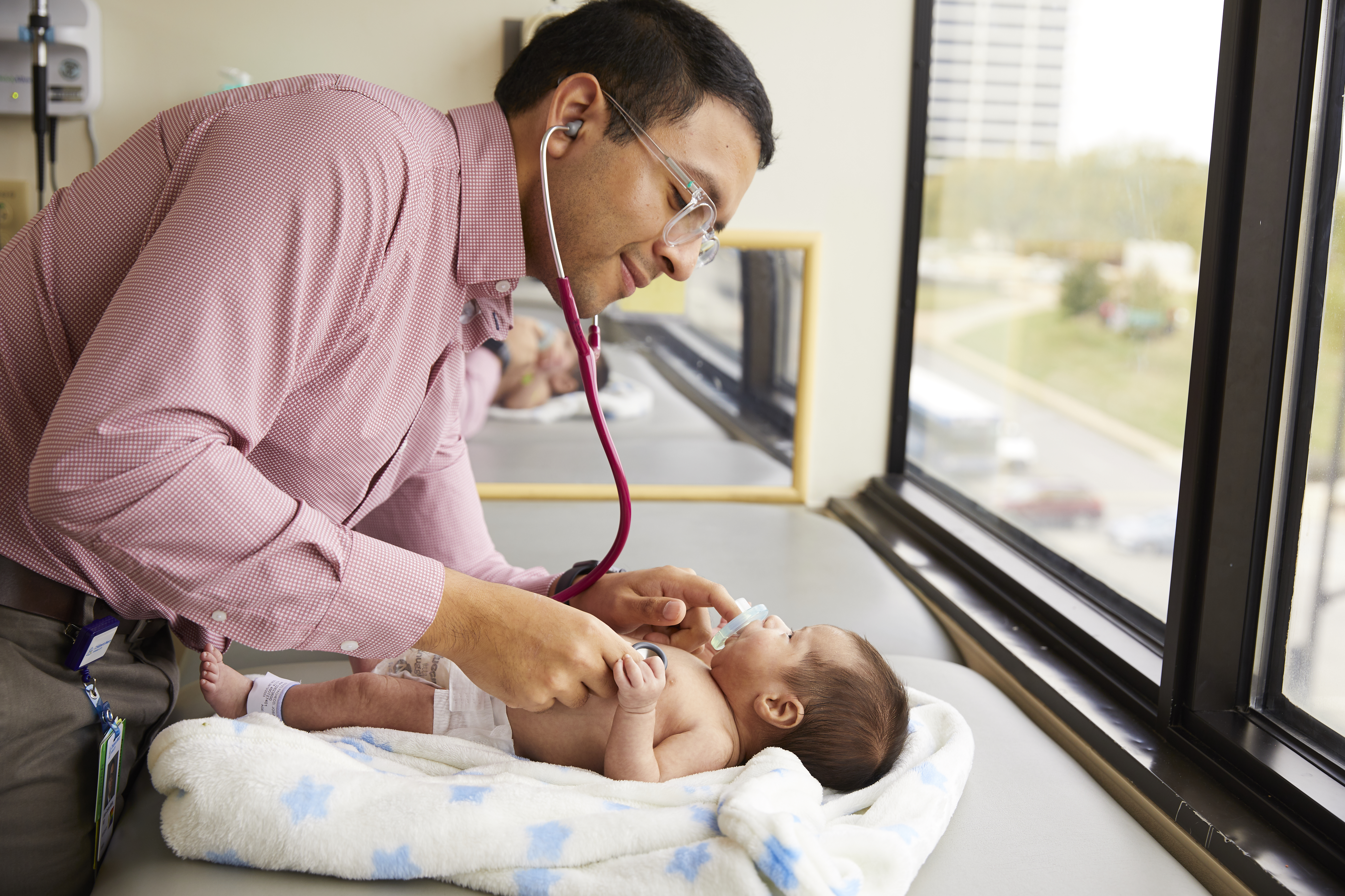 A clinician uses a stethoscope to examine an infant lying on a blanket beside a hospital window.