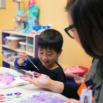 Child painting on an adult's hand during an art activity at a table with paint supplies
