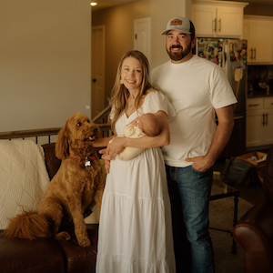 A family portrait taken indoors featuring two adults, an infant, and a brown dog. One adult is holding the baby while standing beside another adult. The dog is sitting on a couch next to them. The photo is set in a warmly lit living room with kitchen cabinets visible in the background.