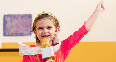 Child wearing a tiara and costume, holding a toy microphone and raising one arm during an imaginative play activity.
