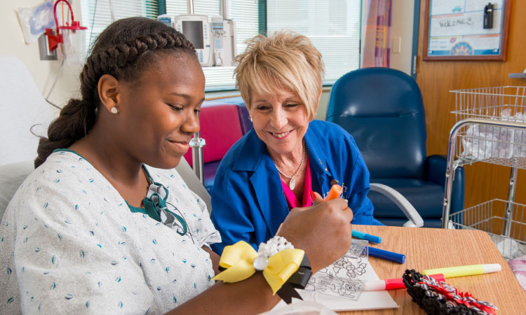 Patient seated at a hospital bedside table coloring a picture with markers while a health care team member sits nearby and engages in the activity.