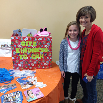 Child and adult standing beside a display table featuring a decorated collection box with the message 'Give Kindness to CM' and materials promoting donations and acts of kindness for Children's Mercy.