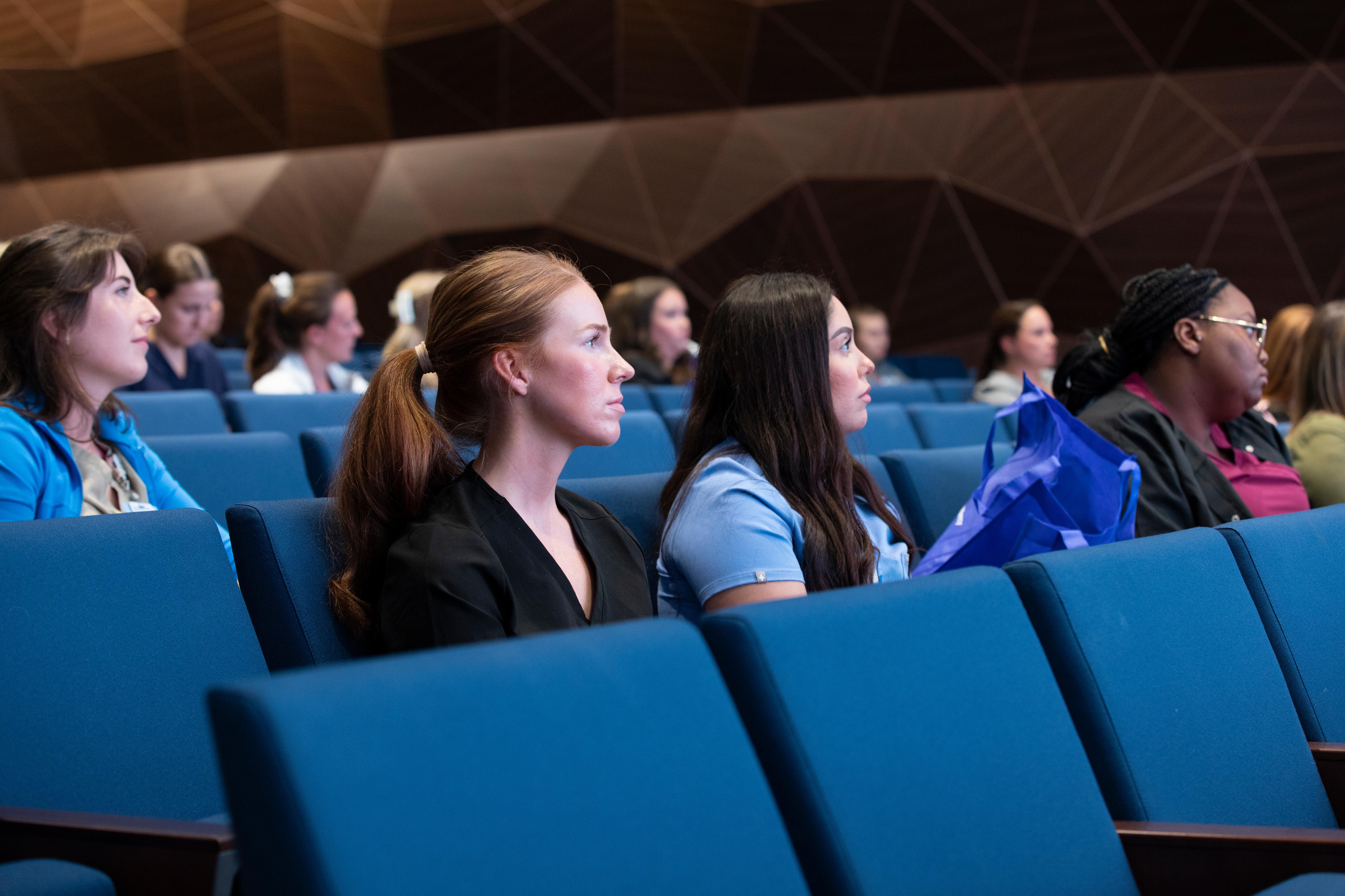 photo of employees sitting in blue chairs in an auditorium looking forward at a speaker