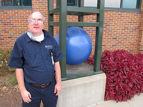 Person standing outdoors beside a large blue spherical sculpture in a metal frame outside a brick building.
