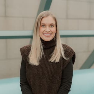 Professional portrait of a person with shoulder-length hair wearing a dark sleeveless knit turtleneck over a long-sleeve top. The person is standing indoors in front of a light-colored architectural background and looking toward the camera.