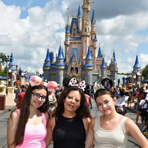 Three people posing in front of a large castle at a theme park. Two people are wearing character-themed headbands. The castle and a busy walkway with park visitors are visible in the background under a partly cloudy sky.