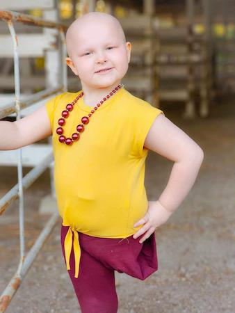 Child wearing a bright yellow top and beaded necklace standing outdoors beside a metal railing.