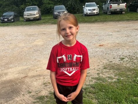 A young girl with a blond ponytail and a red sports uniform smiles in a gravel parking lot.