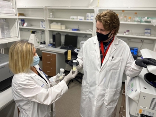 Staci Elliott, Clinical Research Coordinator in Neonatology, hands off a breast milk sample to Cas LeMaster, Bioinformatics Scientist.  Both are wearing masks over their mouths and wearing white lab coats and gloves. 