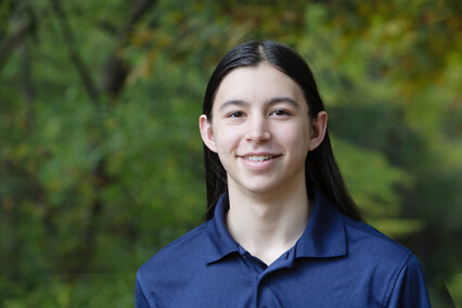 Portrait of a young person wearing a blue collared shirt, photographed outdoors with trees and greenery in the background