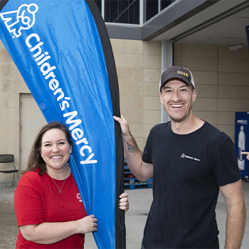 Two people standing outdoors beside a blue Children's Mercy banner at a community event.