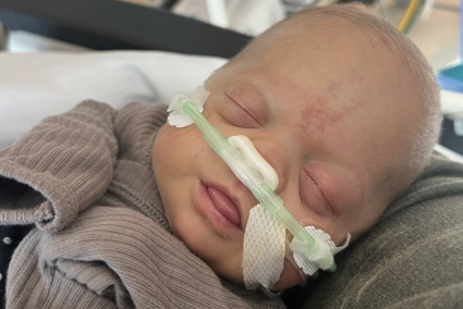 A close-up photo of a newborn baby with closed eyes, wearing a nasal cannula for oxygen support. The baby is dressed in a gray outfit and is lying on a green pillow.