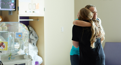Two adults standing and embracing in a hospital room, with a newborn bassinet and patient care equipment visible nearby.