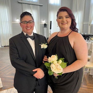 Two people posing indoors at a formal event. One person is wearing a black tuxedo with a bow tie and boutonniere, and the other is wearing a black sleeveless dress while holding a bouquet of white flowers. The photo was taken in a bright event space with tables and white drapery in the background.
